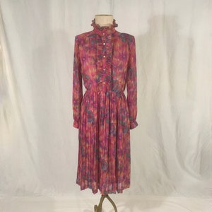 Può includere: Un abito vintage rosa con stampa floreale, maniche lunghe e gonna a pieghe. L'abito presenta un colletto alto a balze e una fila di bottoni. L'abito è esposto su un manichino in metallo dorato.