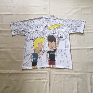 May include: White t-shirt with a cartoon print of two characters from the animated series "Beavis and Butt-Head". The characters are standing in front of a crowd of people. The text "I MEAN, IT'S JUST FRIENDS" is printed in a speech bubble above the characters.