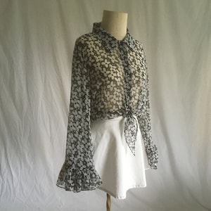 May include: A sheer, long-sleeved blouse with a floral print in black and white. The blouse has a button-up front and a tie at the waist. The sleeves have a ruffled edge.
