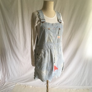 May include: A light blue denim overall dress with white stripes and a red and white patch on the front. The dress is distressed and has a frayed hem.