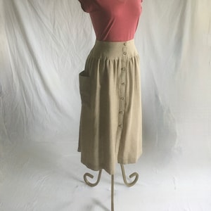 Può includere: Una gonna beige con bottoni e una tasca sul lato sinistro. La gonna è su un manichino che indossa una maglietta rosa polvere a collo a V.