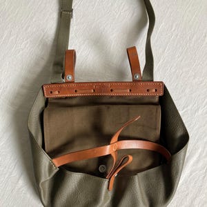 Puede incluir: Un bolso de cuero verde con una correa y hebilla de cuero marrón. El bolso está abierto y el interior está forrado con tela marrón.