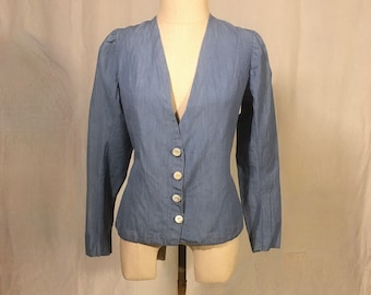 May include: A light blue denim button-up shirt with a deep V-neckline and long sleeves. The shirt is fitted and has a slightly puffed sleeve.
