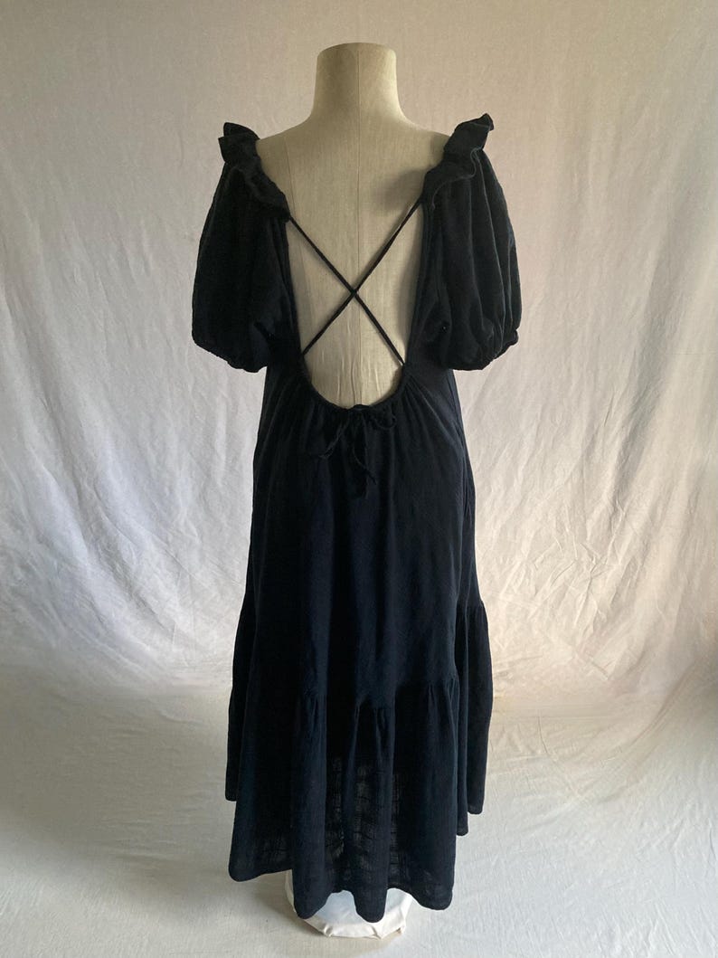 May include: A black maxi dress with a low back and crisscross straps. The dress features short, puffy sleeves and a tiered skirt. The dress is displayed on a mannequin against a white backdrop.