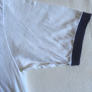May include: White t-shirt with a navy blue ribbed trim around the neck and sleeves.