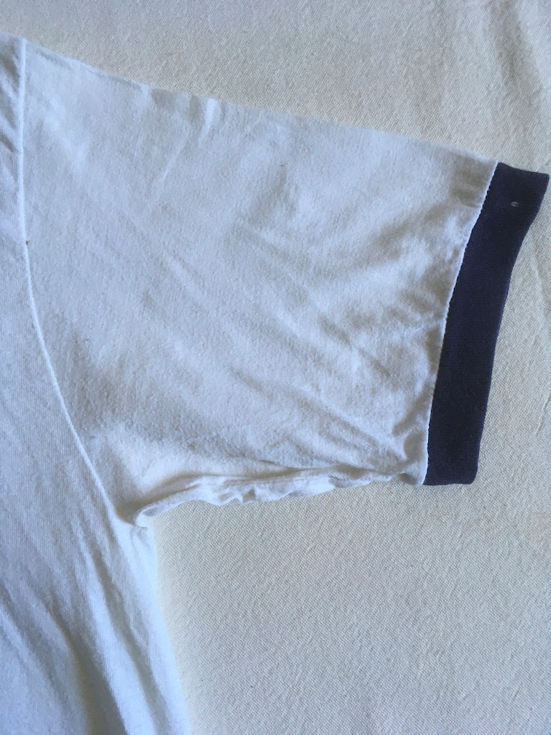 May include: White t-shirt with a navy blue ribbed trim around the neck and sleeves.