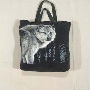 May include: A black canvas tote bag with a white wolf graphic. The wolf is looking to the right and is surrounded by a forest scene.