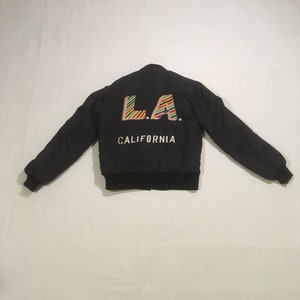 Op de afbeelding: Zwarte bomberjack met een kleurrijke "LA. CALIFORNIA" geborduurde patch op de rug.