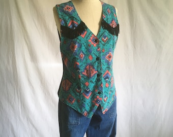 May include: A sleeveless vest with a geometric pattern in shades of blue, green, and pink. The vest has black fringe trim at the neckline and a button closure.