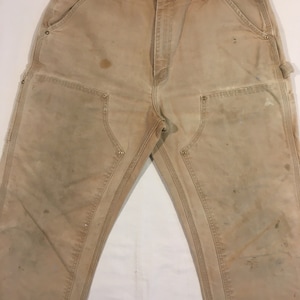 Puede incluir: Un par de pantalones de trabajo de color marr&oacute;n claro con m&uacute;ltiples bolsillos y costuras reforzadas. Los pantalones muestran signos de desgaste, incluyendo manchas y decoloraci&oacute;n.