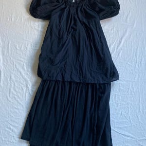 May include: A black dress with an off-the-shoulder neckline and short, puffed sleeves. The dress has a gathered bodice and a tiered skirt, creating a flowing silhouette. The fabric appears to be a lightweight material, suitable for warm weather.