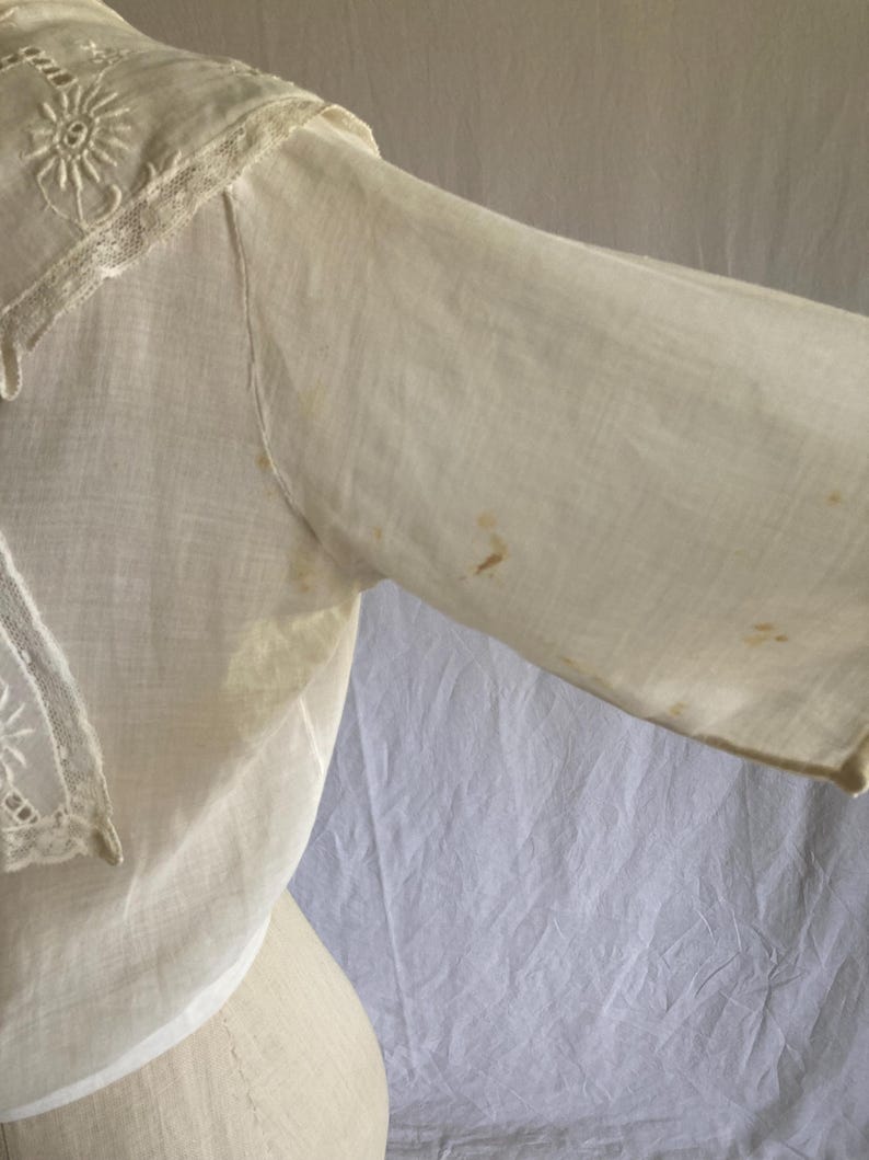 May include: Off-white vintage blouse with a detailed embroidered collar and sleeve. The collar features delicate floral embroidery and lace trim. The sleeve shows some discoloration. The blouse is made of a lightweight fabric.