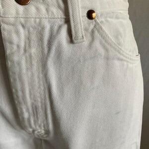 May include: Close-up of white denim jeans with copper-coloured metal buttons and rivets. The jeans feature a classic five-pocket design and a belt loop. The Wrangler brand is visible on the button.