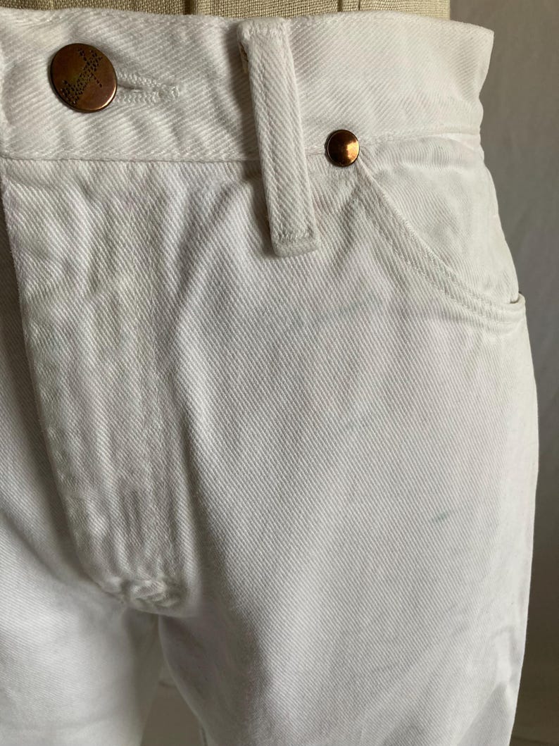 May include: Close-up of white denim jeans with copper-coloured metal buttons and rivets. The jeans feature a classic five-pocket design and a belt loop. The Wrangler brand is visible on the button.
