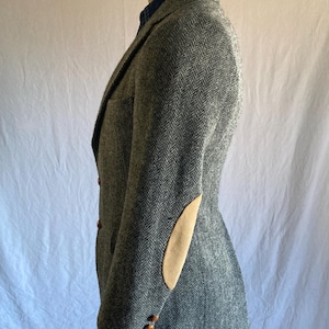 May include: A grey herringbone blazer featuring tan suede elbow patches, a notched lapel, and three brown buttons on the sleeve cuffs. The garment is worn over a blue collared shirt and is displayed on a mannequin torso against a plain backdrop.