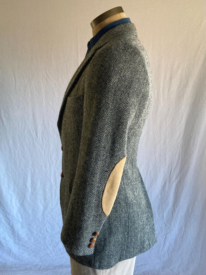 May include: A grey herringbone blazer featuring tan suede elbow patches, a notched lapel, and three brown buttons on the sleeve cuffs. The garment is worn over a blue collared shirt and is displayed on a mannequin torso against a plain backdrop.