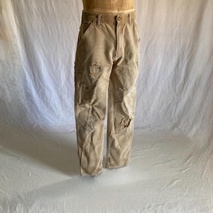 May include: Tan work pants with multiple pockets and distressed details, including a tear on the right knee. The pants are displayed on a mannequin and appear worn.