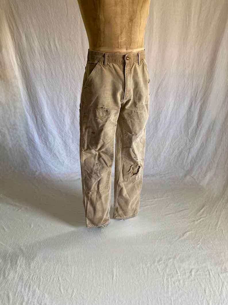 May include: Tan work pants with multiple pockets and distressed details, including a tear on the right knee. The pants are displayed on a mannequin and appear worn.