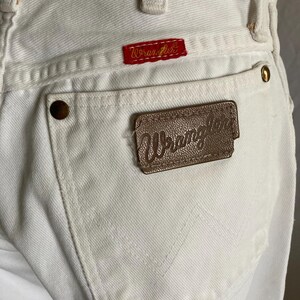 May include: Close-up of white denim jeans with the Wrangler logo on a red patch and a brown leather patch. The jeans have a classic five-pocket design with brass rivets and stitched back pocket detail.