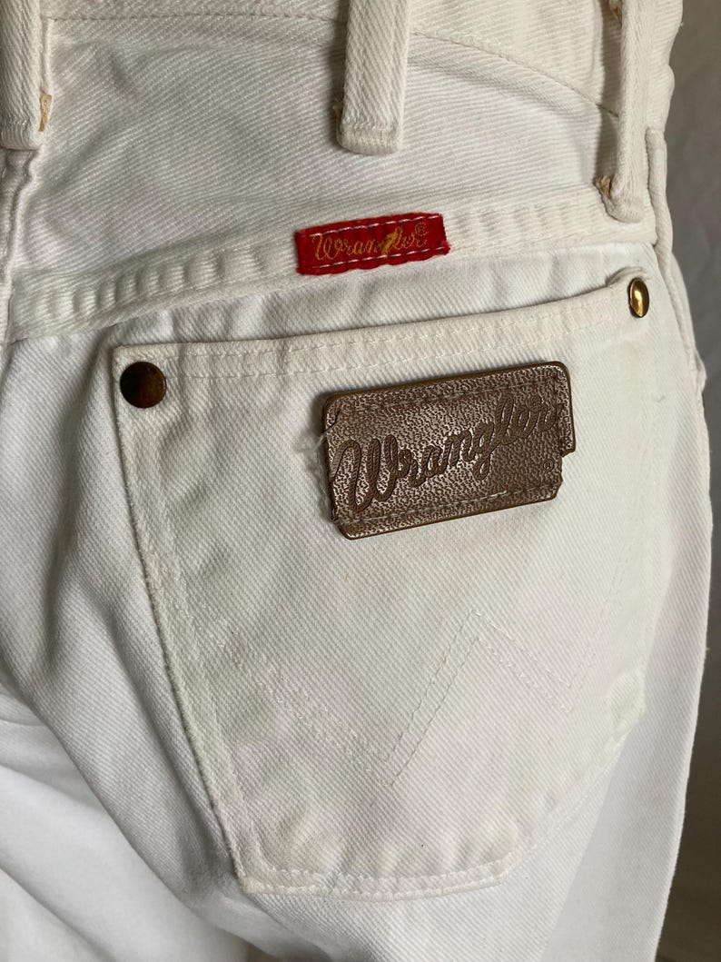 May include: Close-up of white denim jeans with the Wrangler logo on a red patch and a brown leather patch. The jeans have a classic five-pocket design with brass rivets and stitched back pocket detail.