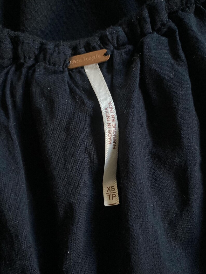 May include: Close-up of a dark navy blue garment with a tag that reads "Free People." A white tag with the text "MADE IN INDIA FABRIQUE EN INDE" and size "XS TP" is attached to the garment.