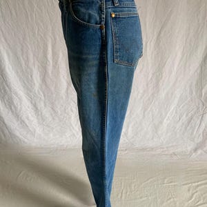 Pu&ograve; includere: Jeans blu in denim con un classico design a cinque tasche. I jeans hanno un taglio dritto e presentano rivetti color ottone e una chiusura con bottone. Il tessuto mostra una sottile sbiaditura e usura, suggerendo un'estetica vintage. I jeans sono esposti su un manichino.