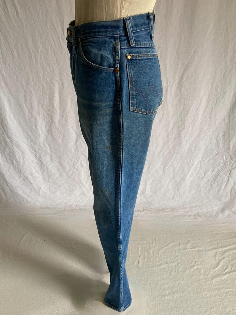 Pu&ograve; includere: Jeans blu in denim con un classico design a cinque tasche. I jeans hanno un taglio dritto e presentano rivetti color ottone e una chiusura con bottone. Il tessuto mostra una sottile sbiaditura e usura, suggerendo un'estetica vintage. I jeans sono esposti su un manichino.