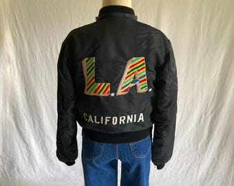 May include: A black bomber jacket featuring the letters L.A. on the back in a multi-colored diagonal striped pattern with a tan outline. Below the letters, the word CALIFORNIA is embroidered in white block lettering. The jacket has a black collar.