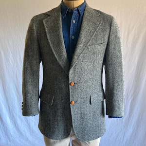 May include: A gray and black herringbone blazer featuring two brown textured buttons, a notched lapel, and two flap pockets. The blazer is worn over a dark blue button-down shirt and light-colored corduroy trousers on a mannequin.