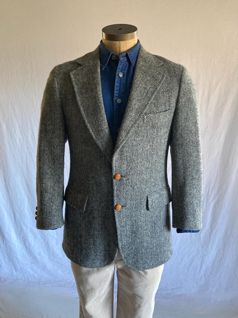 May include: A gray and black herringbone blazer featuring two brown textured buttons, a notched lapel, and two flap pockets. The blazer is worn over a dark blue button-down shirt and light-colored corduroy trousers on a mannequin.