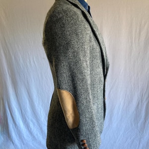 May include: A gray herringbone blazer featuring tan suede elbow patches and three brown buttons on the sleeve cuff. The blazer is worn over a blue collared shirt and is shown from a side profile view, highlighting the textured fabric pattern.