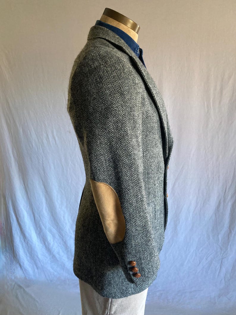 May include: A gray herringbone blazer featuring tan suede elbow patches and three brown buttons on the sleeve cuff. The blazer is worn over a blue collared shirt and is shown from a side profile view, highlighting the textured fabric pattern.