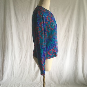 Op de afbeelding: Een turquoise en roze bloemenprint blouse met lange mouwen en een ronde hals.