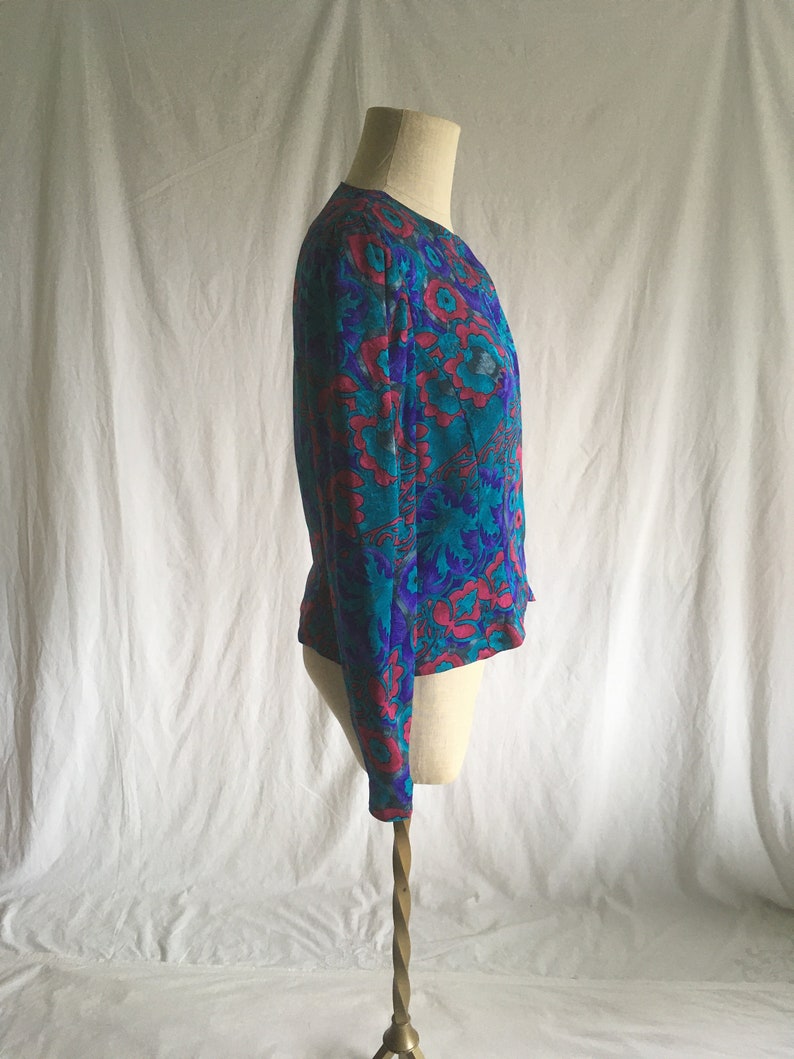 Op de afbeelding: Een turquoise en roze bloemenprint blouse met lange mouwen en een ronde hals.
