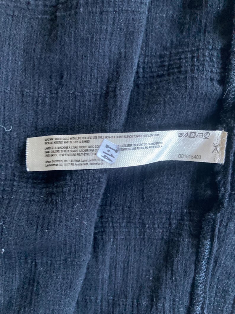 May include: Close-up of a dark blue fabric with a textured pattern. A white tag is attached, displaying washing instructions and a product code. The tag includes text in English and French.