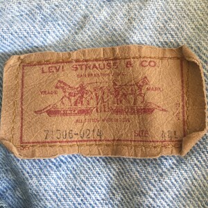 May include: A brown leather Levi Strauss & Co. patch with red stitching. The patch features a red logo with two horses pulling a wagon. The text "Levi Strauss & Co. San Francisco, Cal.  Original  Riveted  Jeans  Mark  All  For  The  Price  Of  One  7306-4214  Size 44L" is printed on the patch.