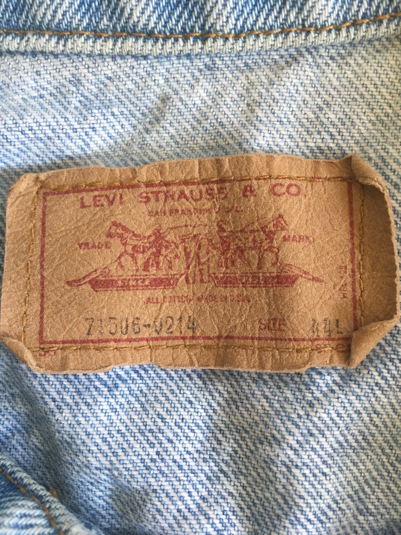 May include: A brown leather Levi Strauss & Co. patch with red stitching. The patch features a red logo with two horses pulling a wagon. The text "Levi Strauss & Co. San Francisco, Cal.  Original  Riveted  Jeans  Mark  All  For  The  Price  Of  One  7306-4214  Size 44L" is printed on the patch.