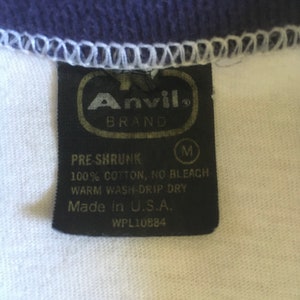 May include: A black and white tag with the text "Anvil Brand" in a gold oval. The tag also includes the text "Pre-Shrunk", "100% Cotton, No Bleach", "Warm Wash-Drip Dry", "Made in U.S.A.", and "WPL10884".