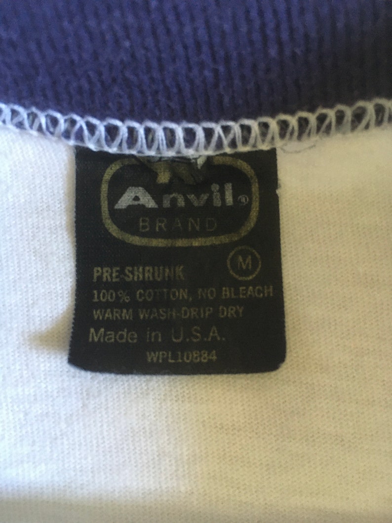 May include: A black and white tag with the text "Anvil Brand" in a gold oval. The tag also includes the text "Pre-Shrunk", "100% Cotton, No Bleach", "Warm Wash-Drip Dry", "Made in U.S.A.", and "WPL10884".