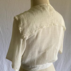 May include: A vintage white blouse with short sleeves, displayed on a mannequin. The blouse features a detailed collar with lace trim and embroidered accents. The fabric appears lightweight and semi-sheer, suggesting a delicate, possibly antique garment.