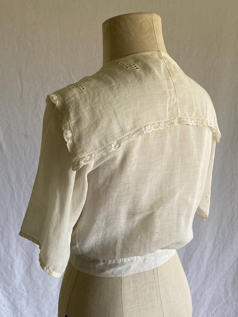 May include: A vintage white blouse with short sleeves, displayed on a mannequin. The blouse features a detailed collar with lace trim and embroidered accents. The fabric appears lightweight and semi-sheer, suggesting a delicate, possibly antique garment.