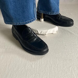 May include: Black leather penny loafers. These classic shoes feature a rounded toe, a strap across the vamp, and a stitched sole. They are shown with blue denim jeans.