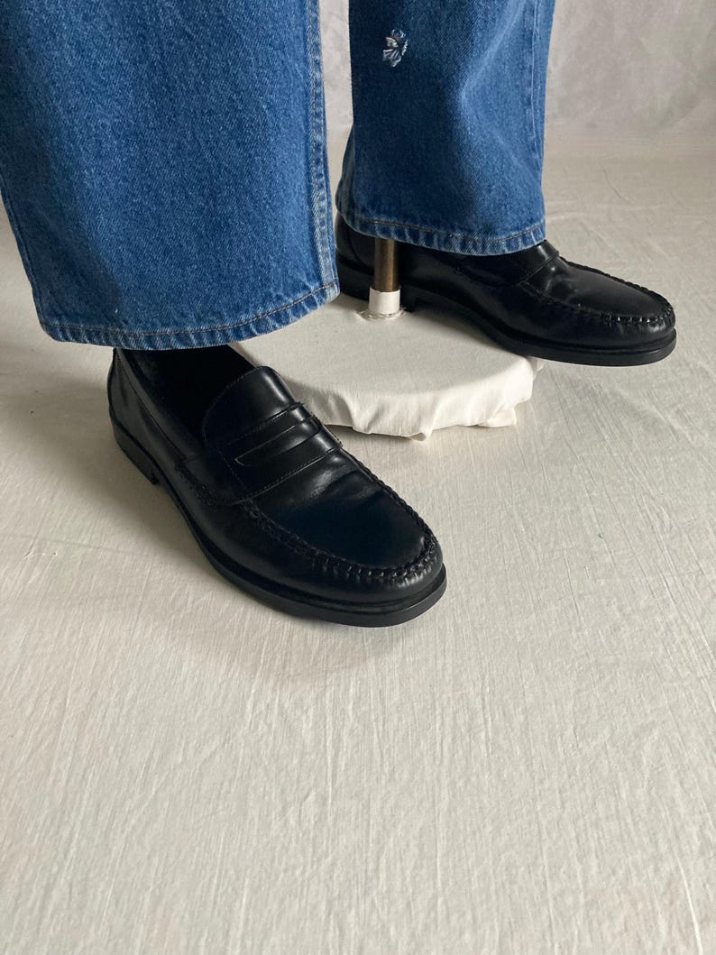 May include: Black leather penny loafers. These classic shoes feature a rounded toe, a strap across the vamp, and a stitched sole. They are shown with blue denim jeans.