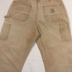 Puede incluir: Un pantal&oacute;n de trabajo de color beige con varios bolsillos y un parche con el logotipo de Carhartt en el bolsillo trasero derecho.