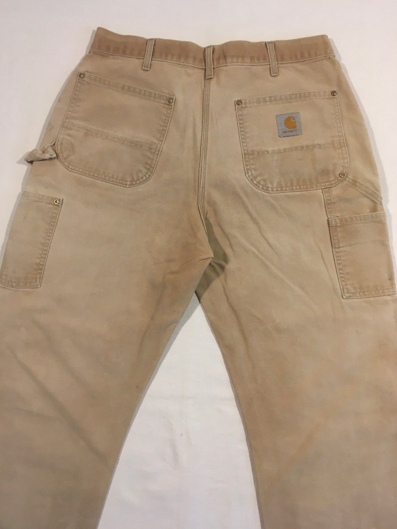 Puede incluir: Un pantal&oacute;n de trabajo de color beige con varios bolsillos y un parche con el logotipo de Carhartt en el bolsillo trasero derecho.