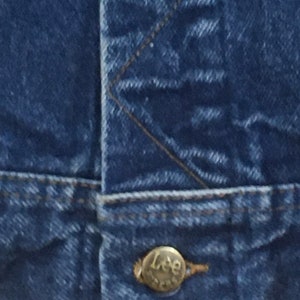 Op de afbeelding: Close-up van een blauw denim jack met een metalen knoop waarop "Lee" staat.