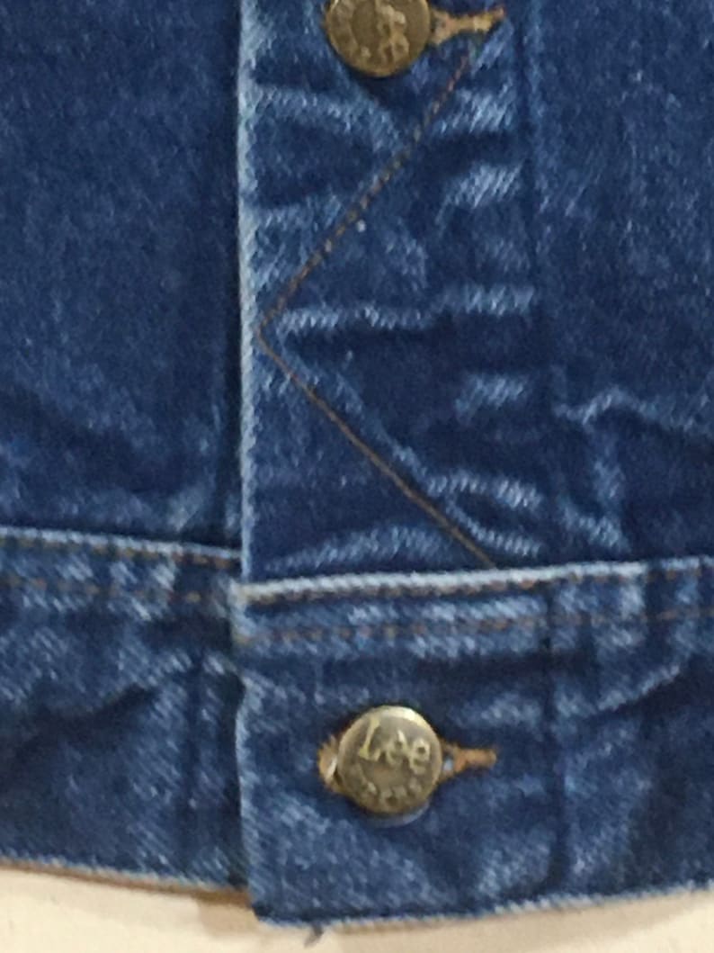 Op de afbeelding: Close-up van een blauw denim jack met een metalen knoop waarop "Lee" staat.