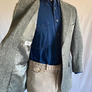 May include: A grey and white herringbone blazer with a grey lining featuring a label that reads The Village Tailor, worn over a dark blue button-down shirt and light beige corduroy trousers on a mannequin torso.