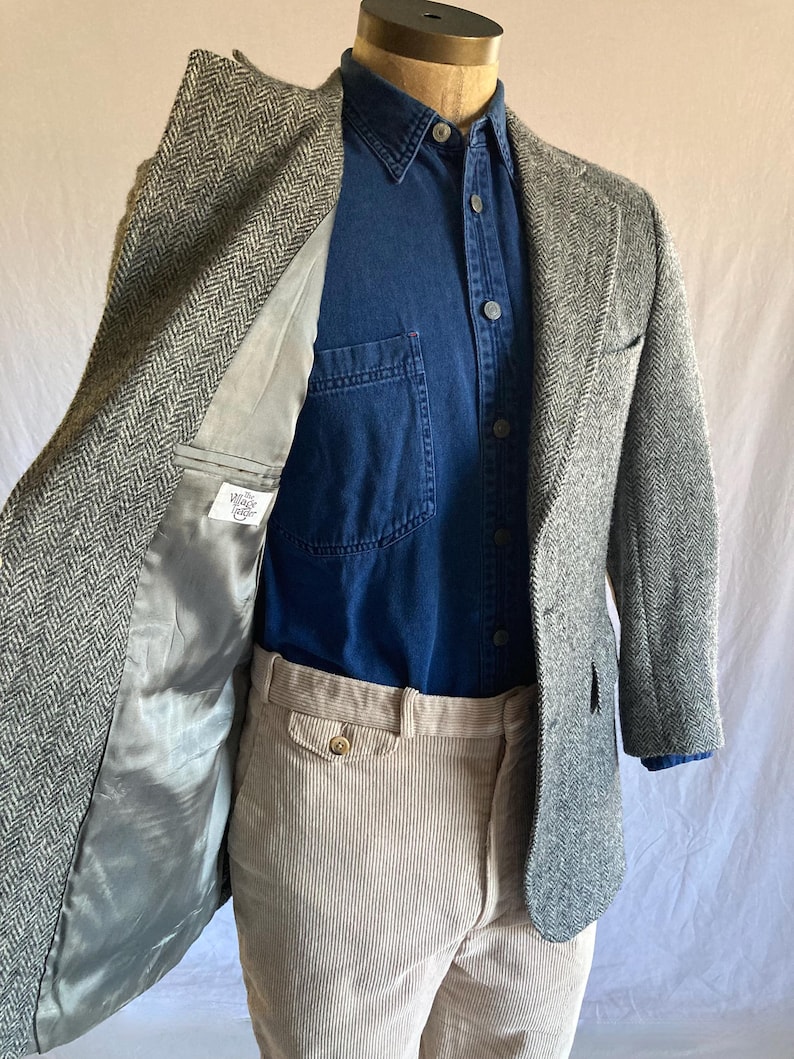 May include: A grey and white herringbone blazer with a grey lining featuring a label that reads The Village Tailor, worn over a dark blue button-down shirt and light beige corduroy trousers on a mannequin torso.