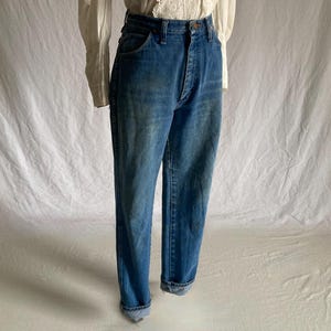 Pu&ograve; includere: Jeans blu in denim con vita alta e chiusura con bottoni. I jeans hanno un classico design a cinque tasche e sono risvoltati alle caviglie. I jeans sono abbinati a una camicetta bianca con dettagli in pizzo.